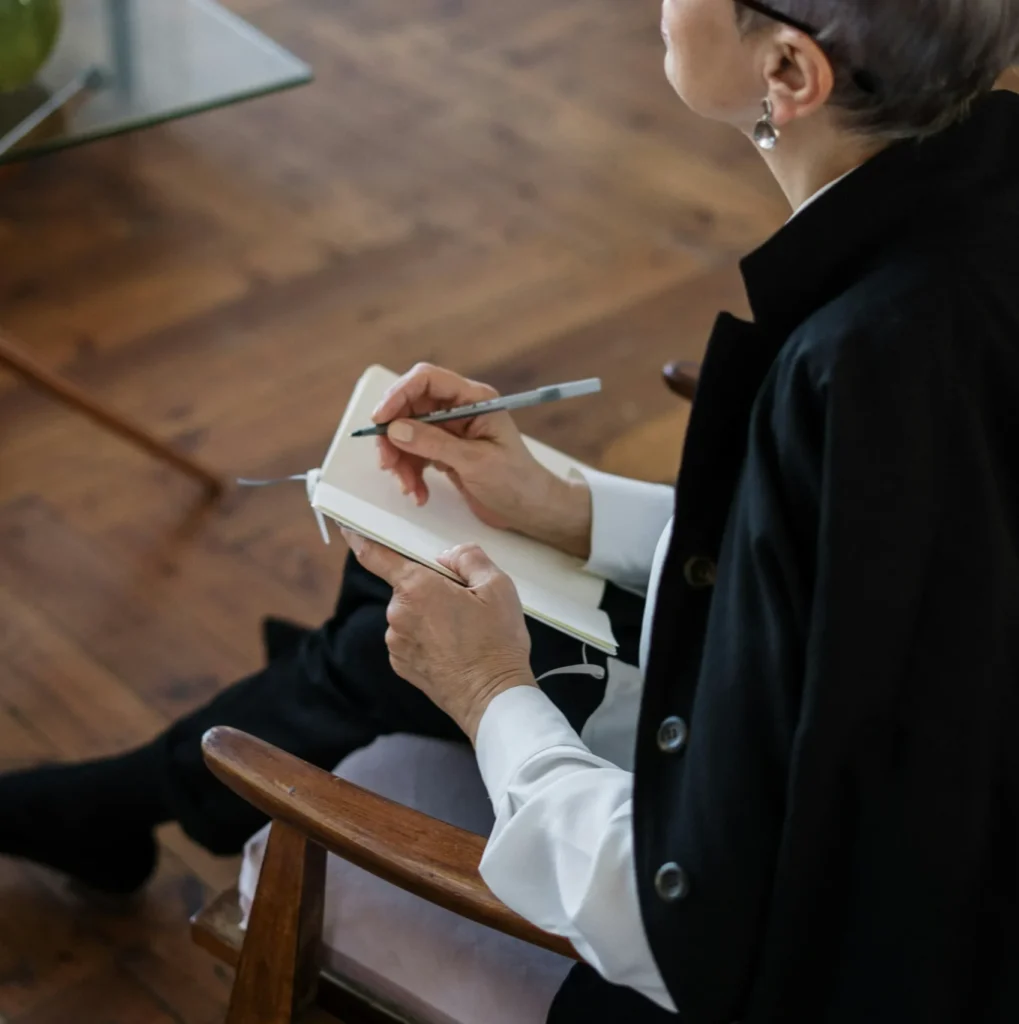 An individual with short gray hair and glasses, wearing a black jacket, sits in a wooden chair and writes in a notebook on their lap. A glass table with a green vase is seen in the background.