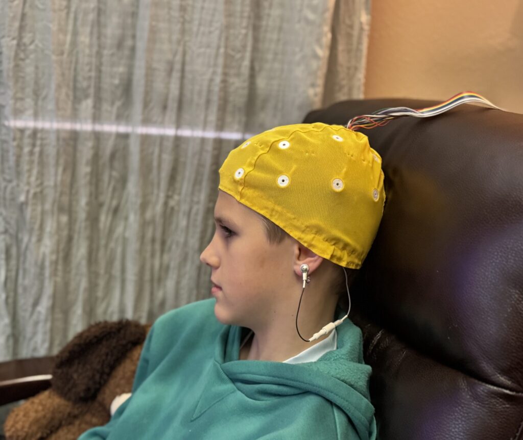 A child sits in a chair wearing a yellow EEG cap with wires attached.